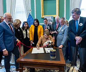 A group of people standing around a table as one person signs a document.