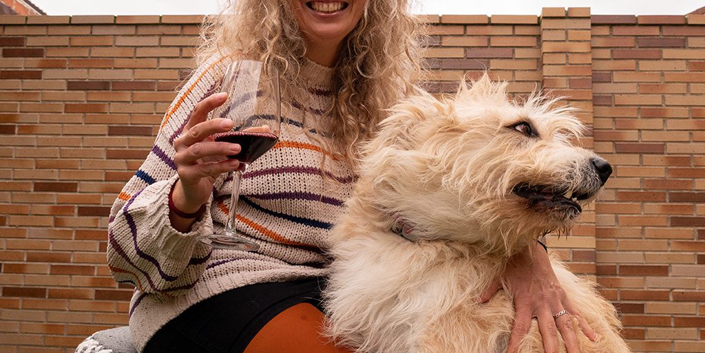 A person smiling, holding a glass of red wine, next to a large white dog.